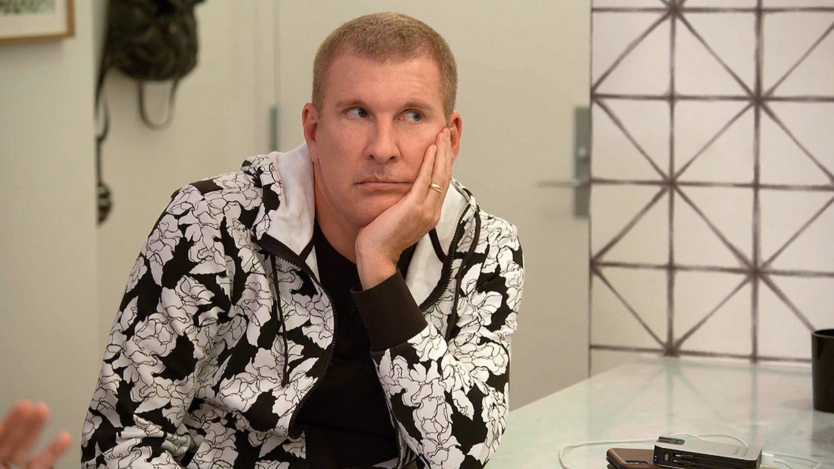 Todd Chrisley looked perplexed in a white and black jacket and black undershirt with his hand on his cheek while filming "Chrisley Knows Best"