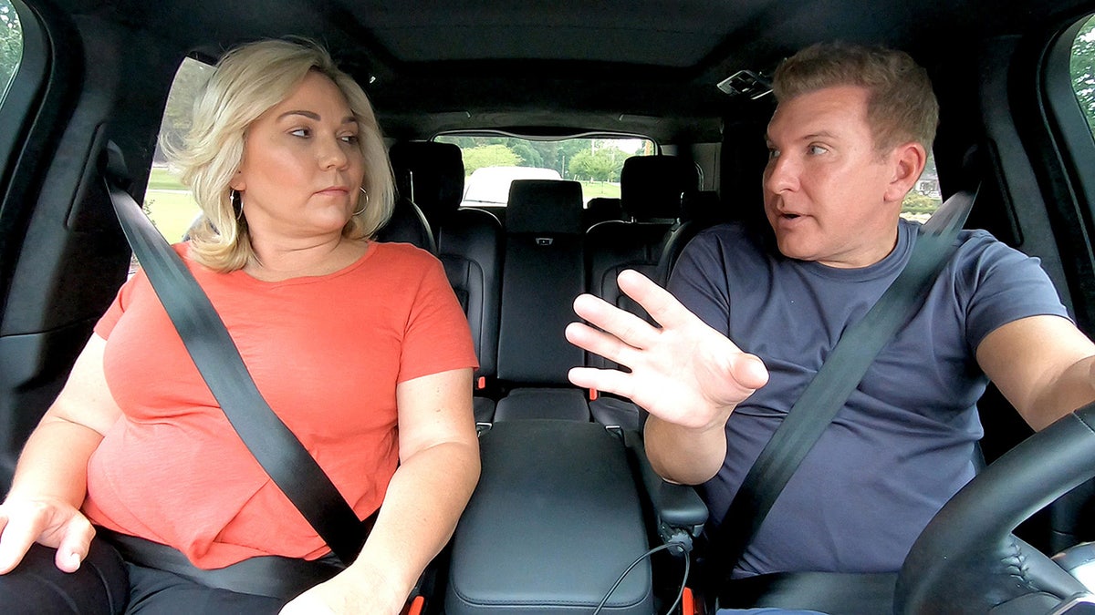 Todd and Julie Chrisley look at each other while Todd drives in a navy long sleeve shirt and Julie wears a bright red short-sleeve shirt filming "Chrisley Know's Best"
