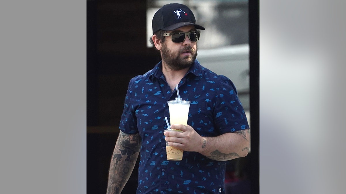 Jack Osbourne in a blue patterned shirt, baseball hat, carrying two drinks one on top of the other