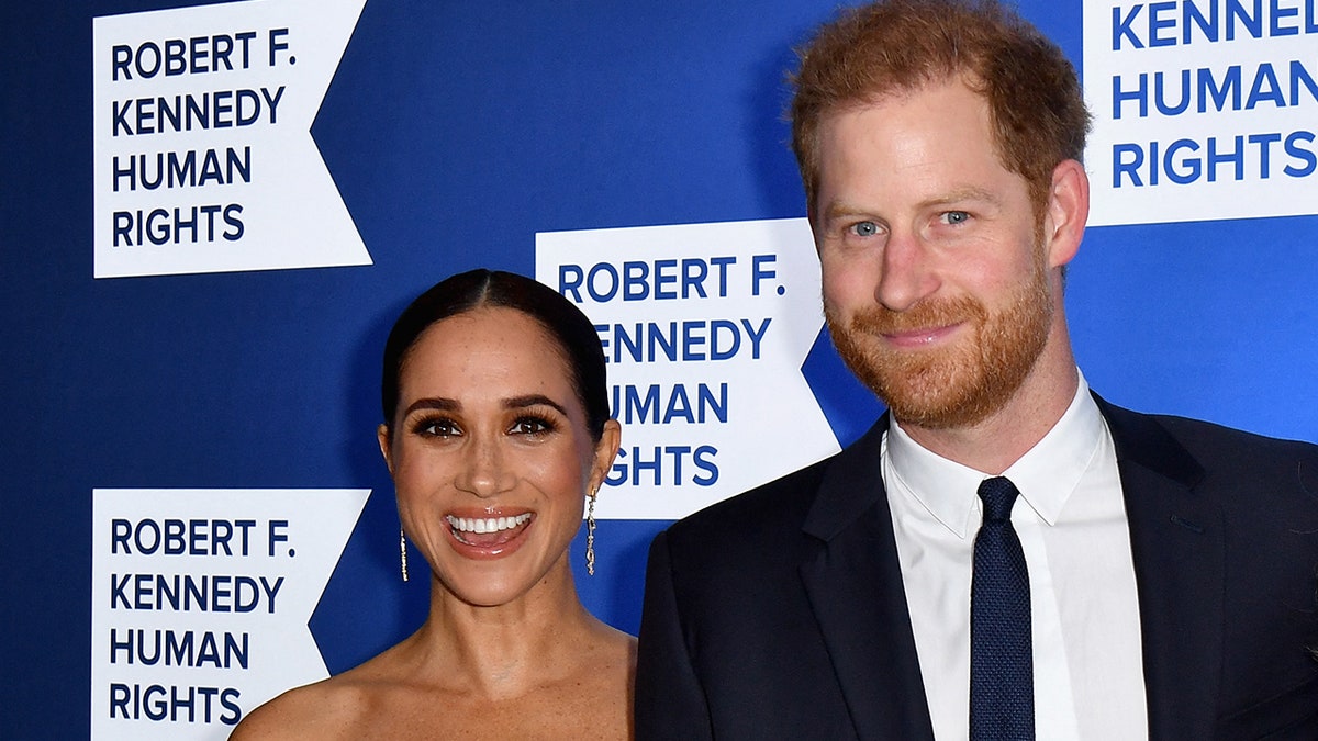 Meghan Markle smiles in a white dress and dangly earrings next to Prince Harry who soft smiles in a navy suit at the Robert F. Kennedy Human Rights Ripple of Hope Award Gala red carpet