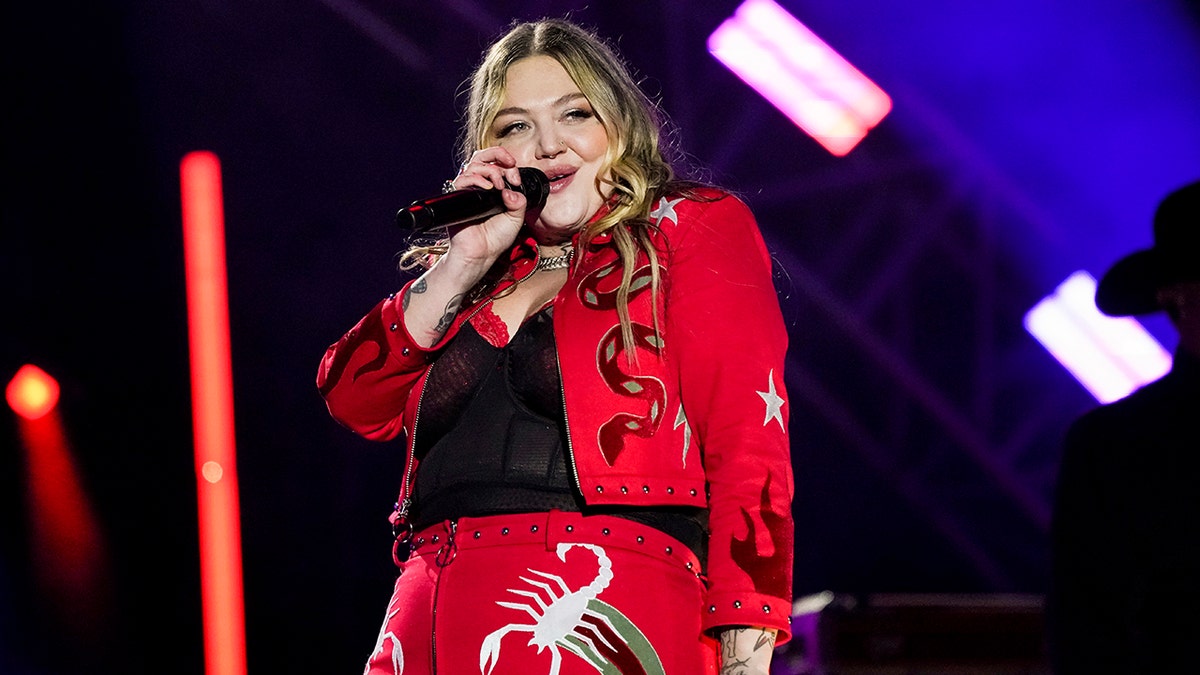 Elle King in a red jacket and pants with snakes and lobsters embroidered, performing on stage
