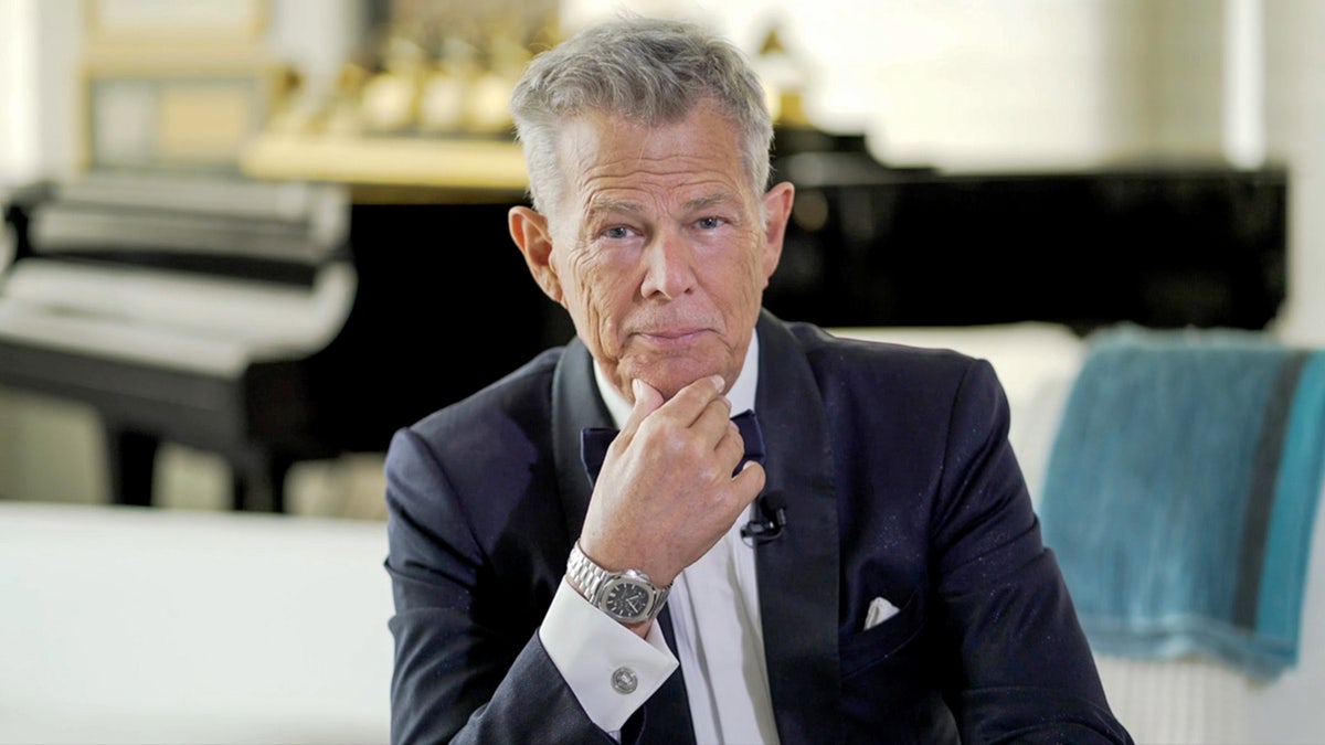 David Foster in a black suit puts his hand on his chin and stares directly at the camera