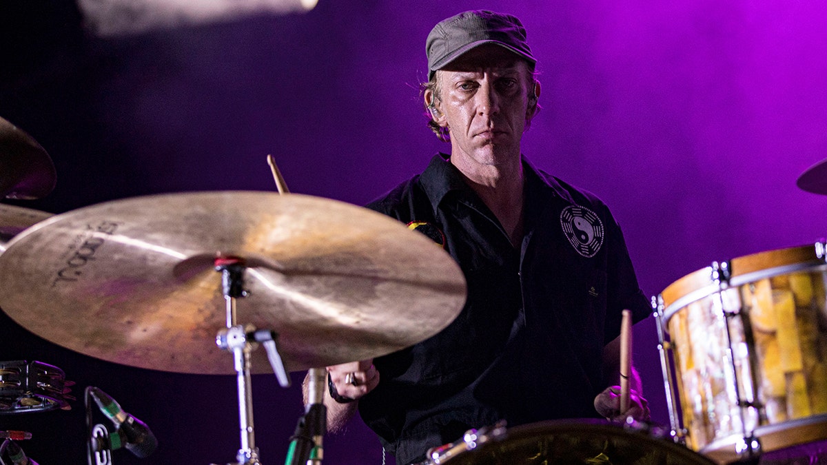 With a purple background Jeremiah Green in a black hat and shirt plays the drums