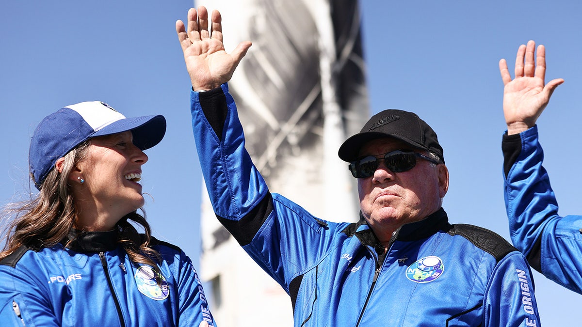 William Shatner in a blue Blue Origin suit and black baseball hat raises his hand to wave to the media after touching down from being in space