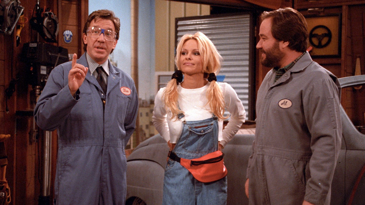 Tim Allen as Tim Taylor in "Home Improvement" in a blue uniform stands next to Pamela Anderson in blue overalls and a white t-shirt and pigtails stands next to Richard Karn as Al Borland also in a dark grey uniform