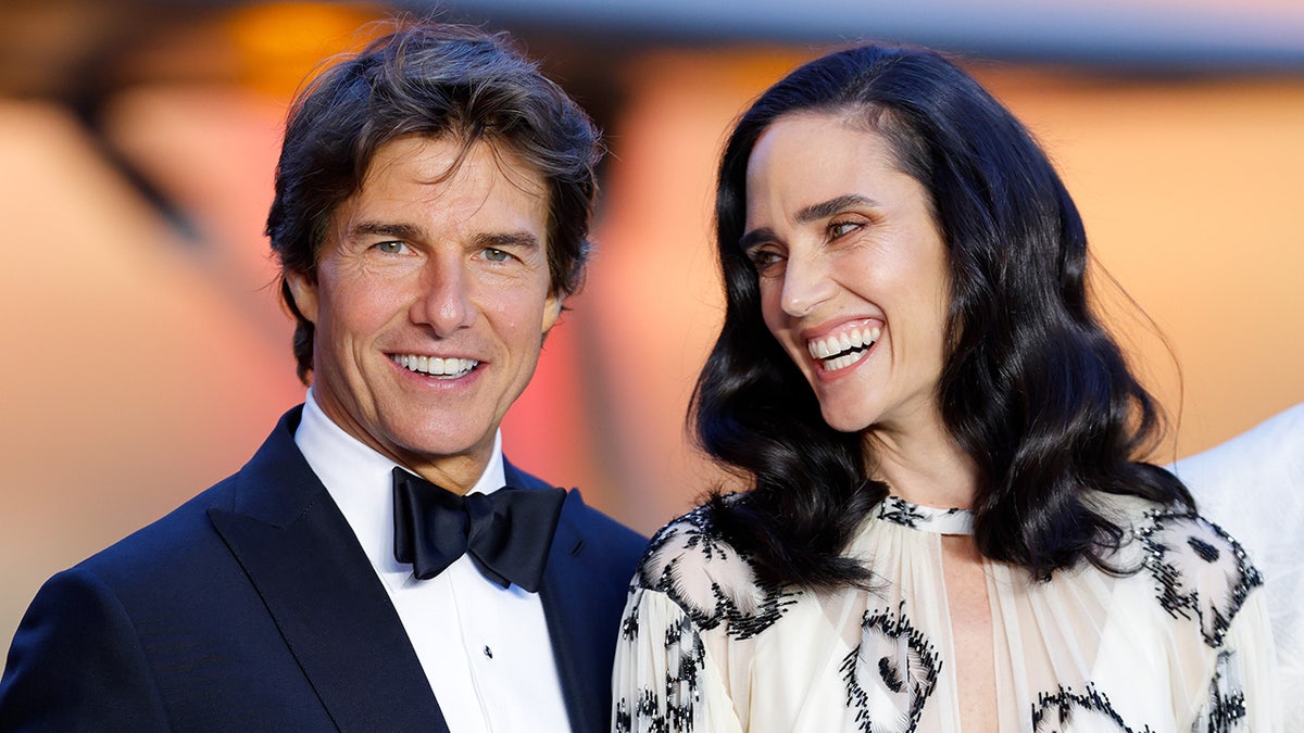 Tom Cruise smiles at the camera in a traditional tuxedo while Jennifer Connelly smiles in a light yellow with black shapes dress at the London premiere of "Top Gun: Maverick"