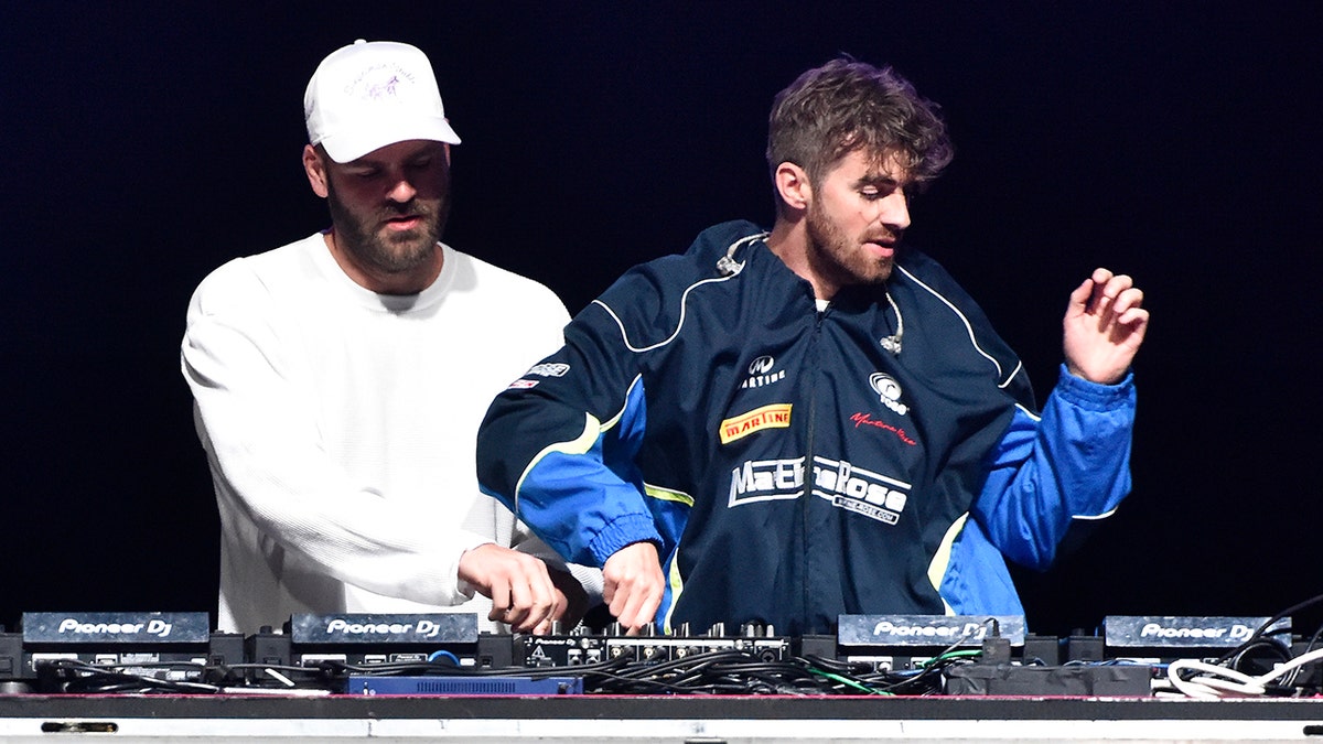 Alex Pall in a white baseball cap and white shirt stands next to Drew Taggart as he spins the DJ turntable while performing in Nevada