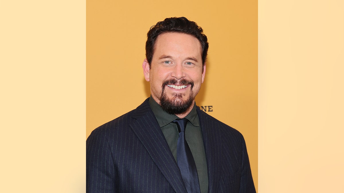 Cole Hauser in front of an orange "Yellowstone" backdrop in a back suit and dark gray shirt