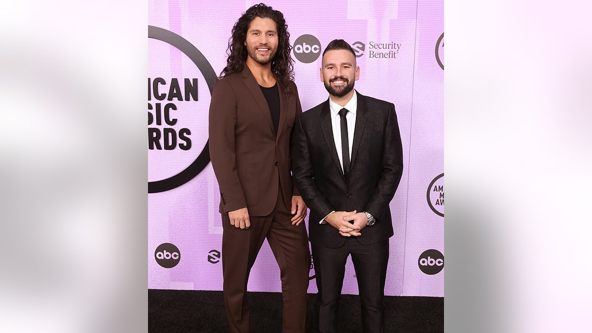 Dan + Shay pose on the carpet at the American Music Awards in a maroon suit (Dan) and black suit (Shay)