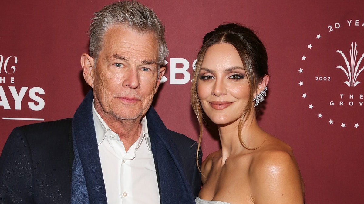 David Foster in a white shirt and dark jacket poses alongside his wife Katharine on the red carpet, she wears her hair in an up-do with two dangly pieces falling forward