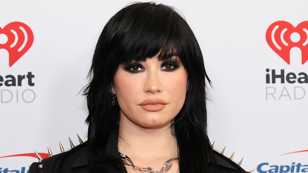 Demi Lovato with black hair and a black jacket with dark eye makeup on the iHeartRadio red carpet in New York City