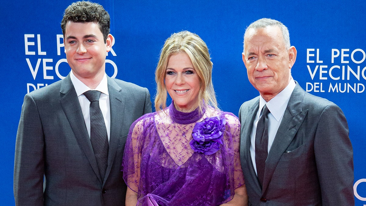 Truman Hanks in a dark suit and tie poses on the red carpet with his mother Rita Wilson in a purple gown with a floral accent and father Tom Hanks in a dark suit