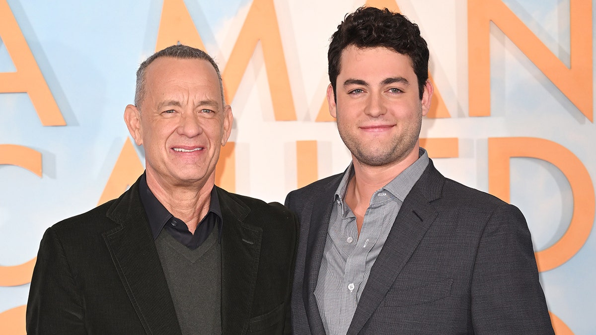 Tom Hanks in a black suit smiles on the red carpet next to his son Truman in a light gray shirt and suit