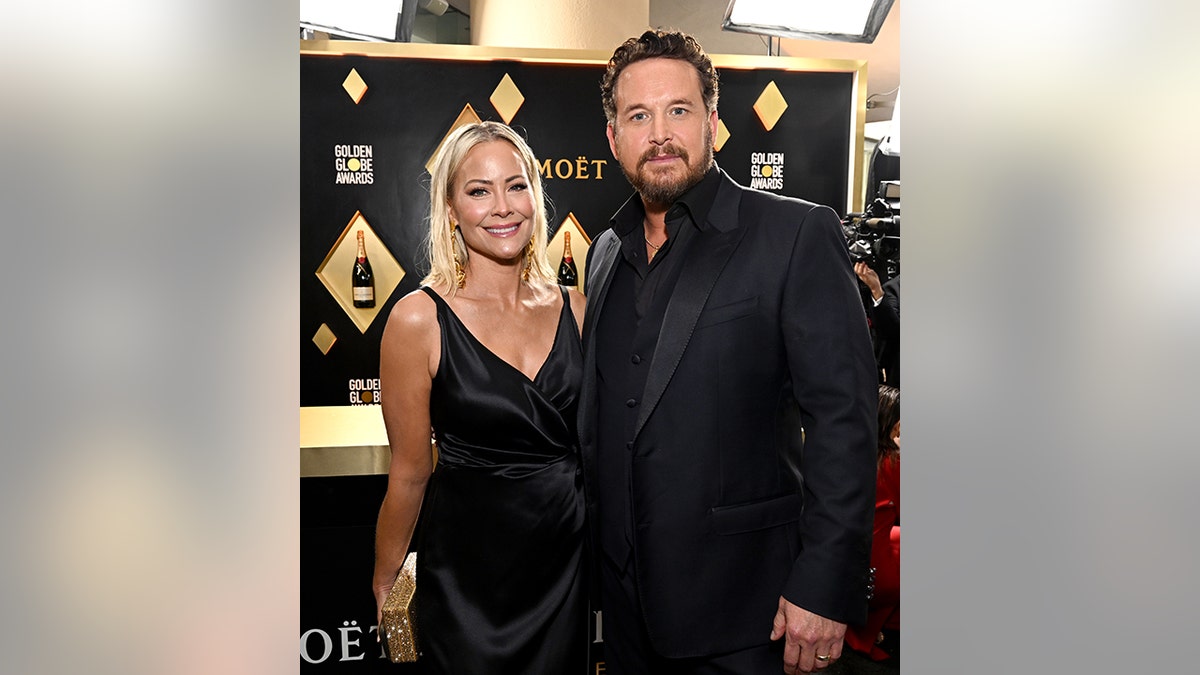 Cole Hauser in a black shirt and tux jacket stands on the Golden Globes red carpet with his wife Cynthia in a black dress