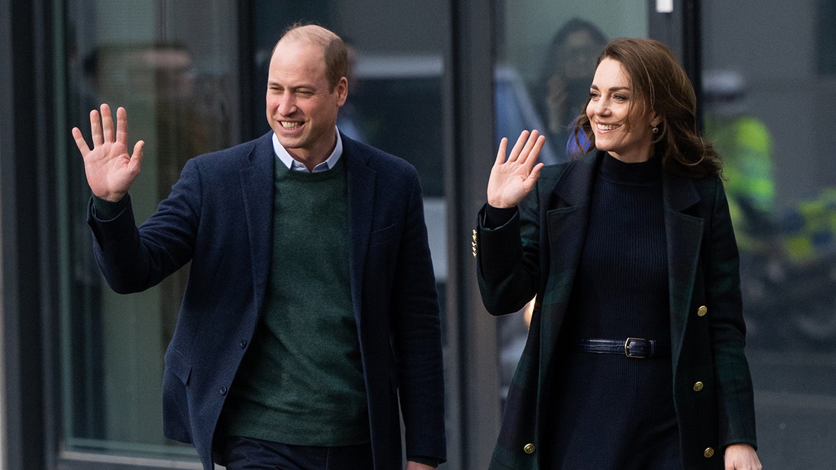 Prince William in a green sweater and blue blazer waves and walks alongside Kate Middleton in a green and blue sweater blazer in Liverpool