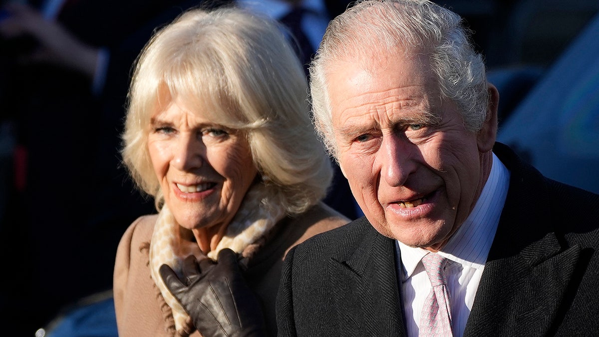 Camilla, the Queen Consort in camel colored jacket walks alongside King Charles in a black suit and pink tie in Bolton