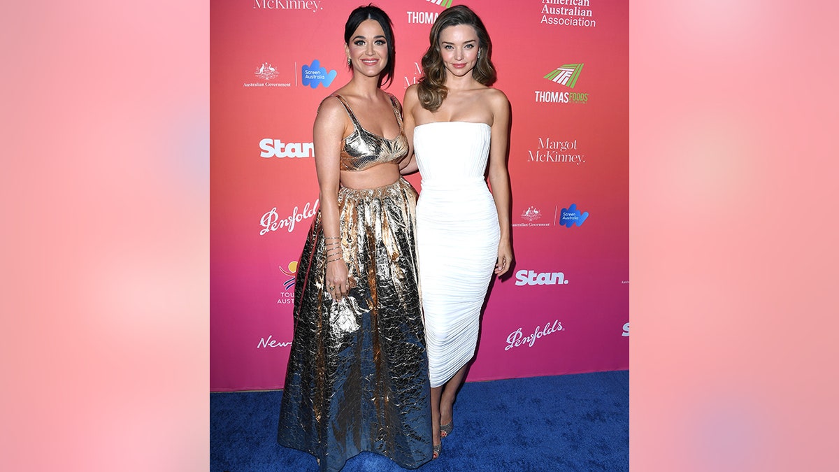 Katy Perry smiles at the camera in a metallic two peace next to Miranda Kerr in an ankle length tight white dress on the red carpet