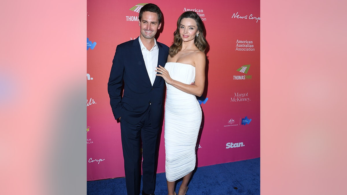 Evan Spiegel in a blue suit jacket and pants with a white button down smiles on the red carpet next to wife Miranda Kerr who places her hand on his chest in a long white tight dress