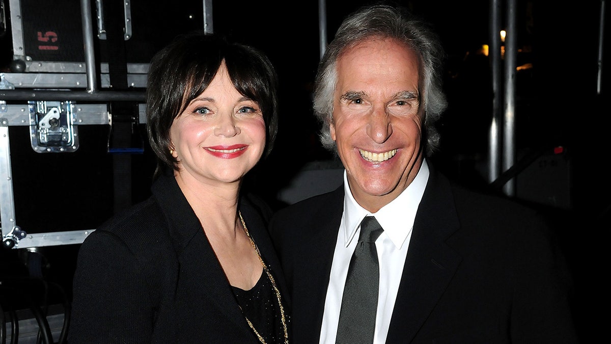 Cindy Williams with red lipstick and a black outfit smiles next to Henry Winkler in a black suit and patterned gray tie