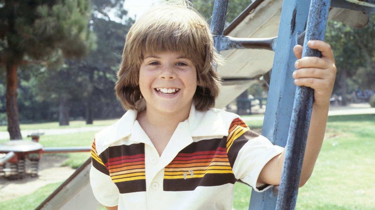 Adam Rich in a white shirt with colored stripes on the top with long floppy hair on "Eight Is Enough"