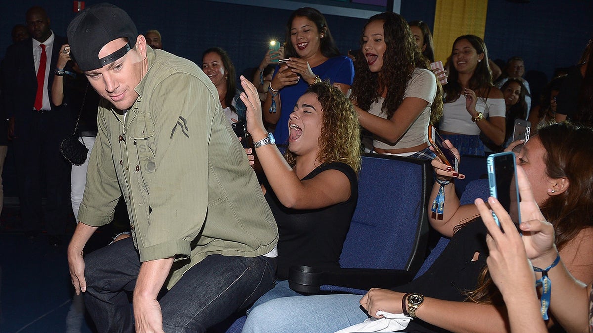 Channing Tatum in an olive green shirt and jeans gives a fan in a chair a little lap dance with his back turned to her