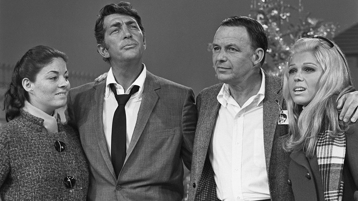 In a black and white photo, Deana Martin stands next to Dean Martin (her father) in a suit, who stands next to Frank Sinatra in a suit with no tie and his daughter Nancy