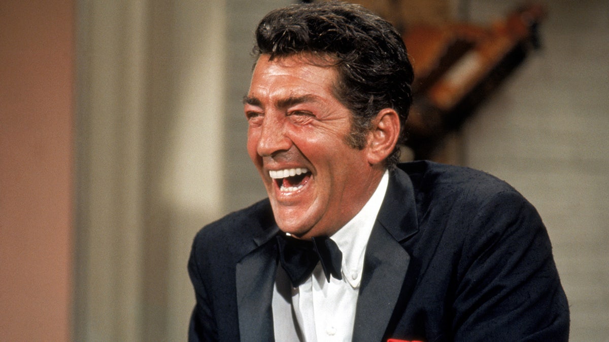 Dean martin in a velvet tuxedo laughs on set of his variety show in 1967