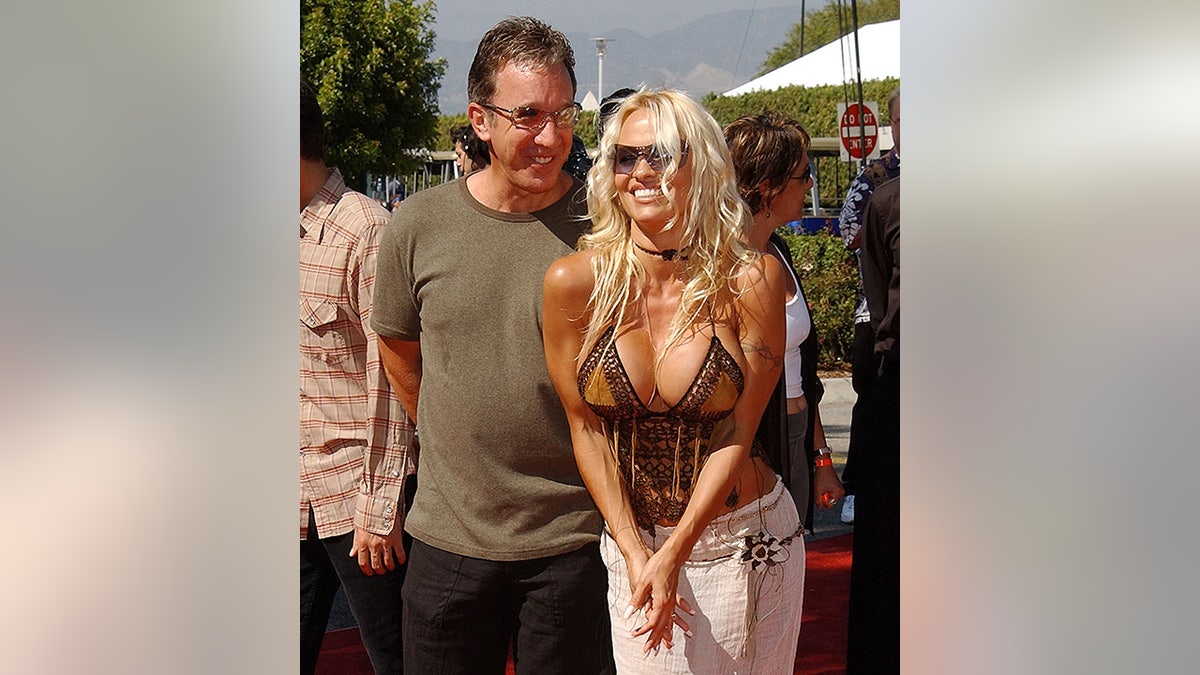 Tim Allen in an olive green t-shirt smiles behind Pamela Anderson in a low-cut tank top and white pants on the 2022 Teen Choice Awards carpet