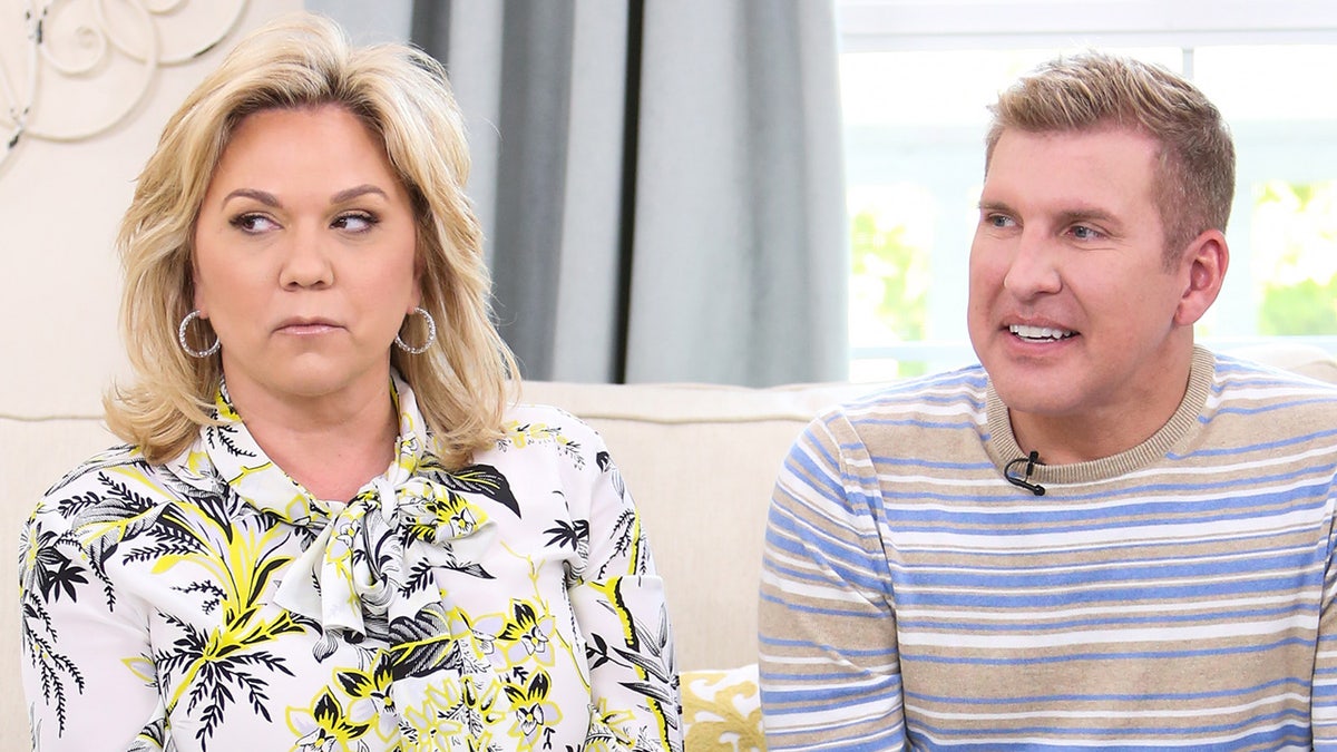 Julie Chrisley looks perplexed in a white floral top sitting on the couch next to a partially smiling Todd Chrisley in a striped shirt