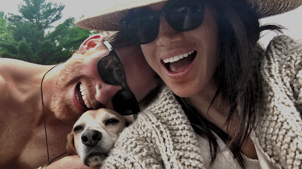 Prince Harry with sunglasses on holds on to his dog with Meghan Markle as she smiles in a crocheted sweater, large brimmed hat and sunglasses