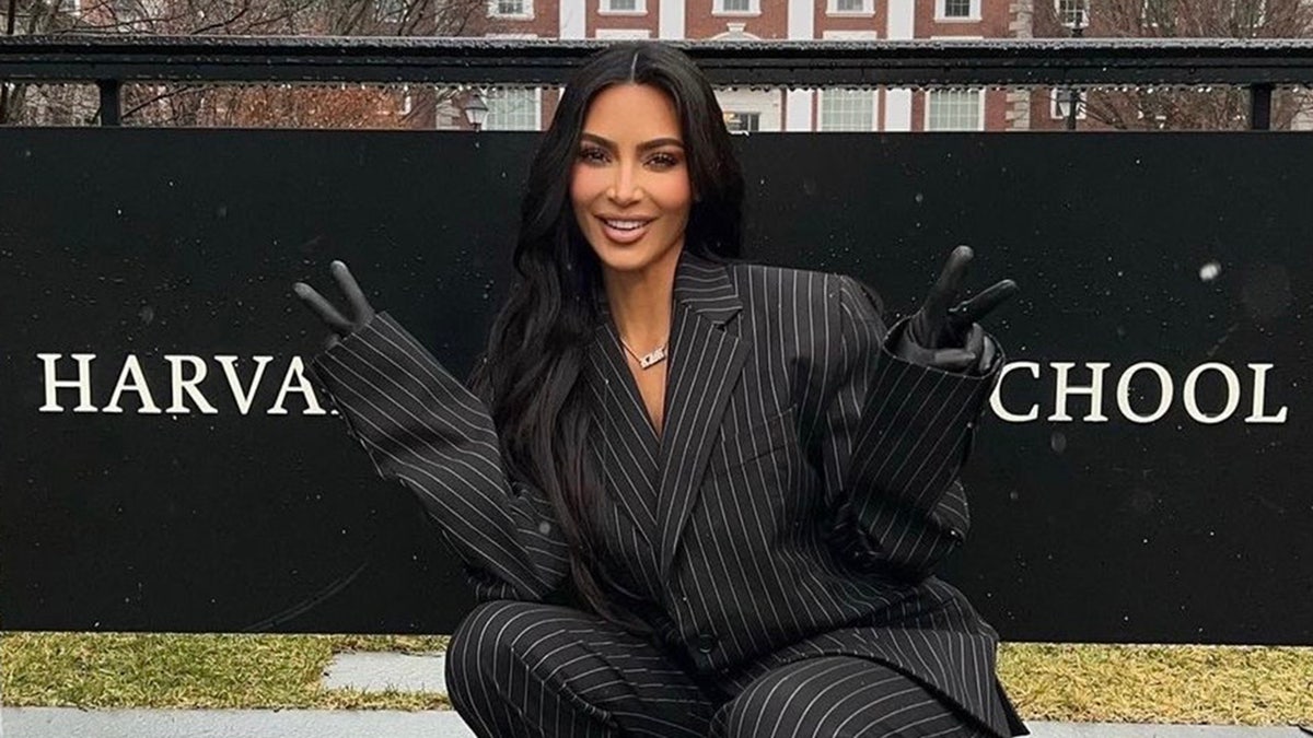 Kim Kardashian em um terno listrado preto e branco faz sinais de paz em frente a uma placa que diz 'Harvard Business School'