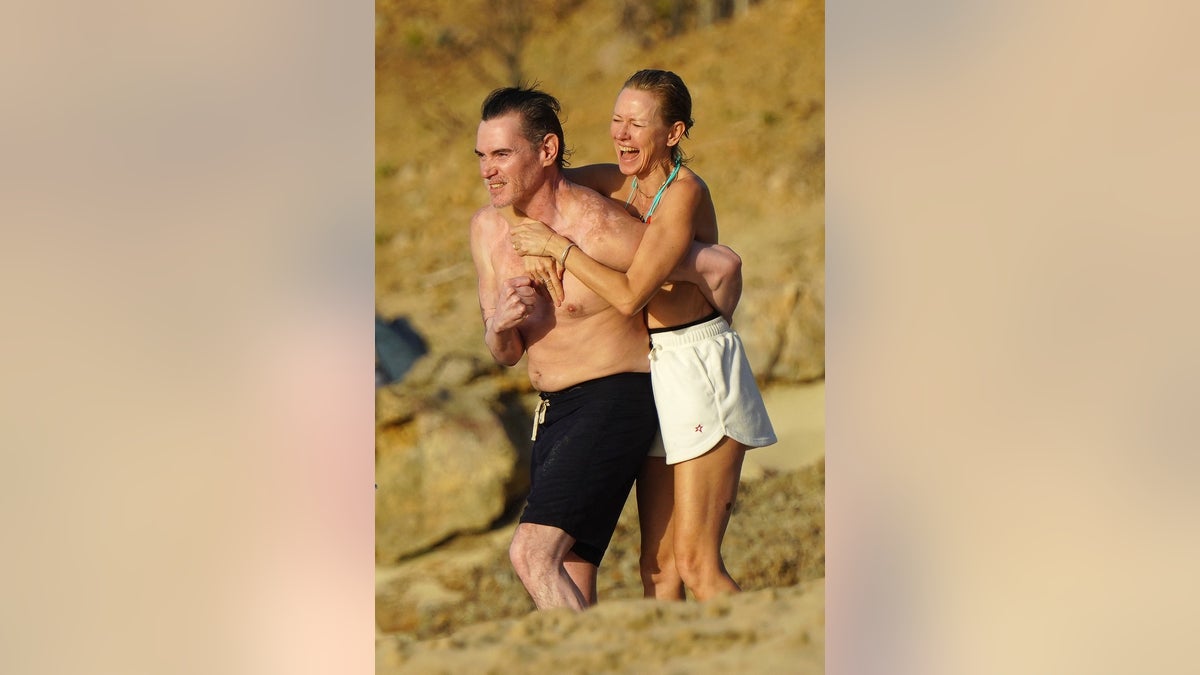 Naomi Watts wearing a bikini top and white shorts holds on to boyfriend Billy Crudup's back and laughs on the beach