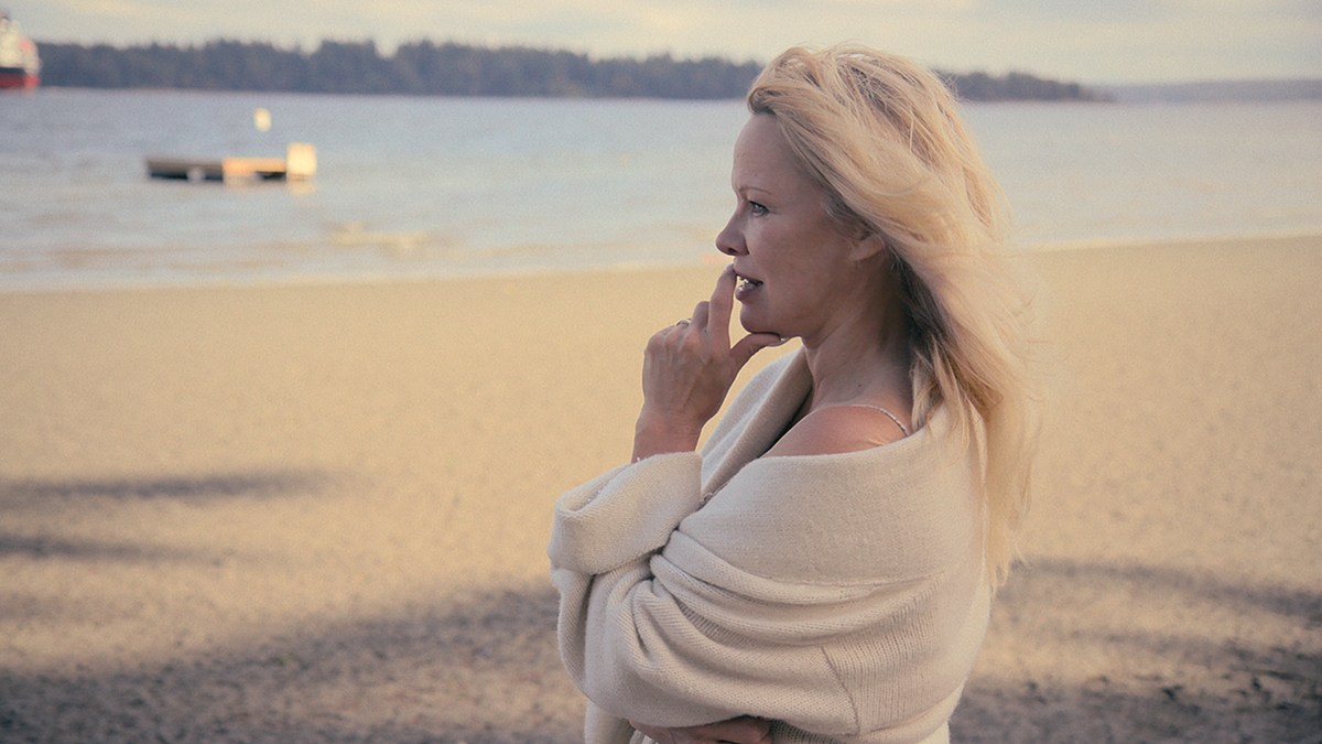 Pamela Anderson wrapped in a cream sweater while she stares off in the distance at the beach