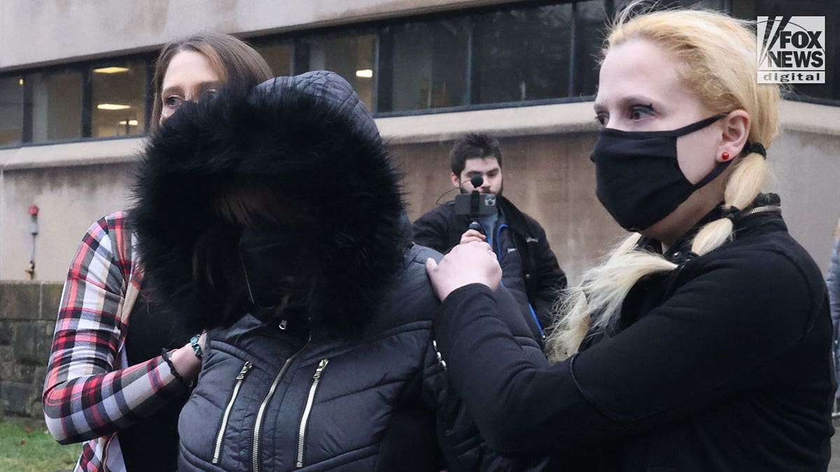 A woman with a black coat on with fur-lined hood pulled over her face is helped to the car by two younger woman holding her shoulders