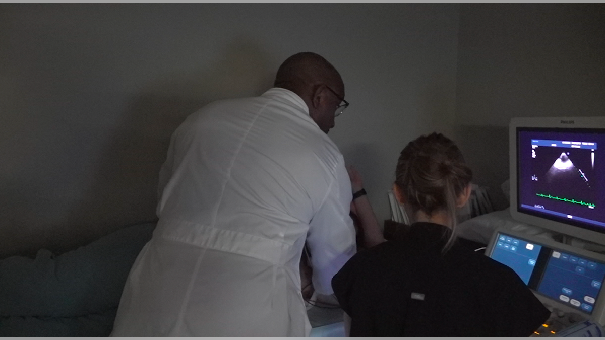 Doctor leaning over a patient standing next to a nurse who is observing the echocardiogram screen