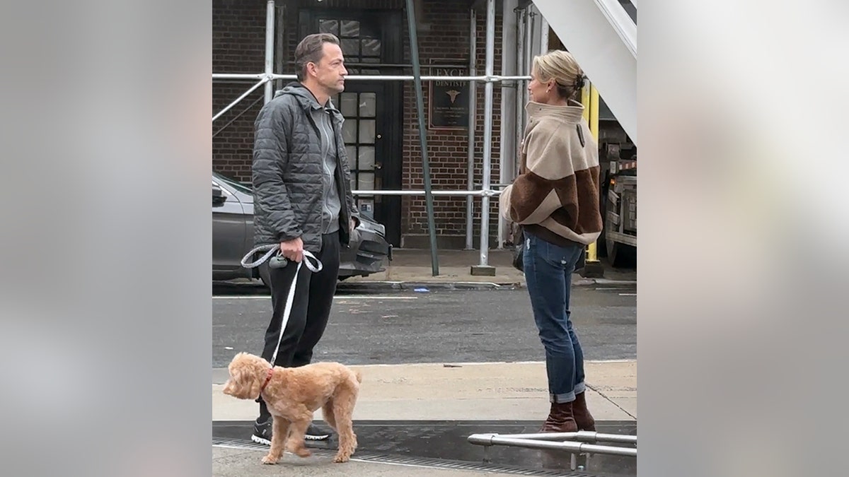 Amy Robach in a two-toned brown coat stands opposite Andrew Shue in a gray coat who holds the leash to dog Brody