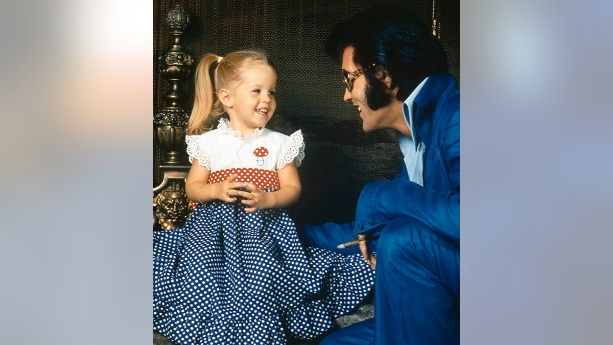 A young Lisa Marie Presley in pigtails and a dress with a white top, red belt, and blue skirt smiles at her father Elvis in a bright blue suit and turquoise shirt