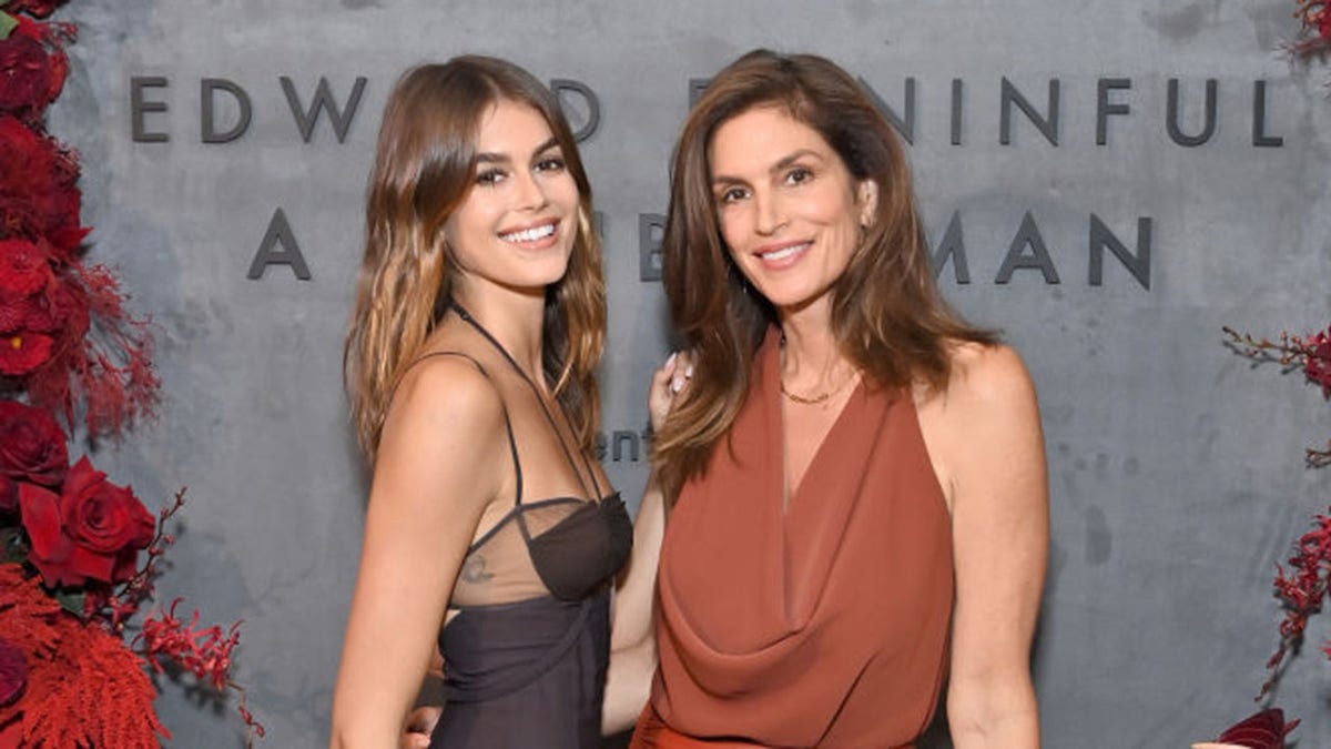Kaia Gerber in a black dress with lace smiles next to her mother Cindy in a burnt orange dress