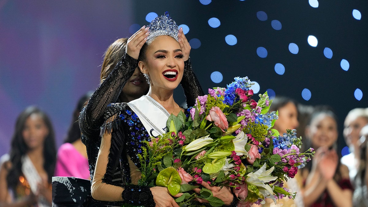 Miss USA R'Bonney Gabriel holds a huge bouquet of flowers as she is crowned Miss Universe on the stage, wearing a sash and blue dress