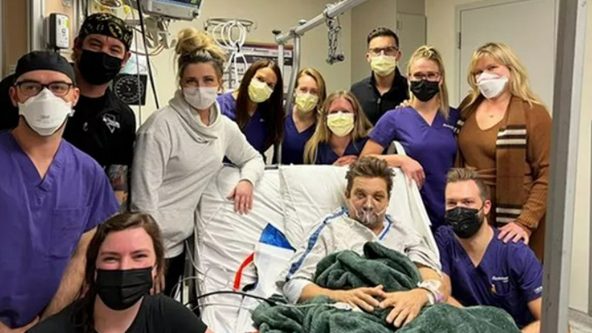 Jeremy Renner lies in a hospital bed with a green blanket and oxygen mask, surrounded by medical staff in the hospital