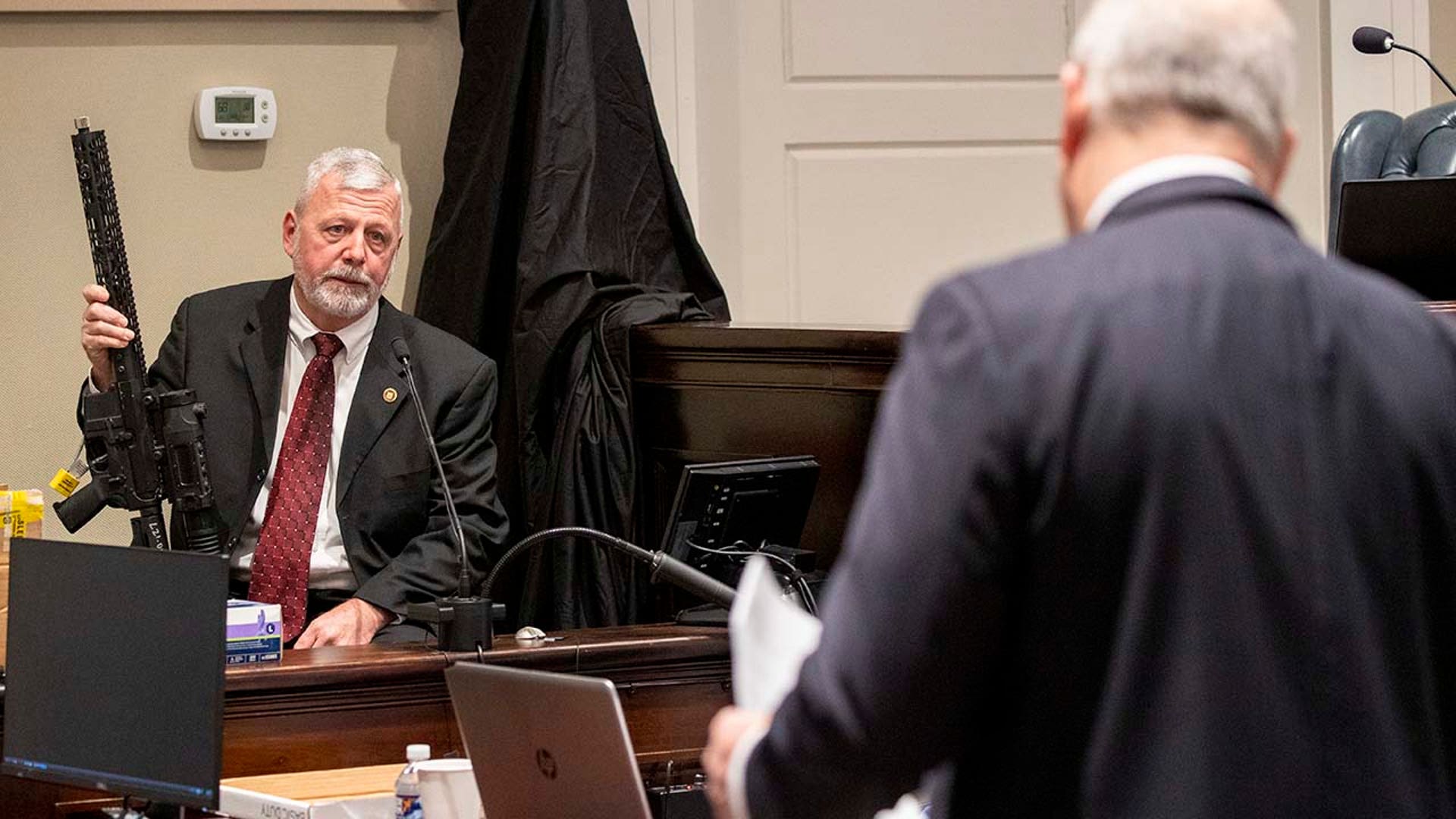 A man holds up a gun while seated at a witness stand.