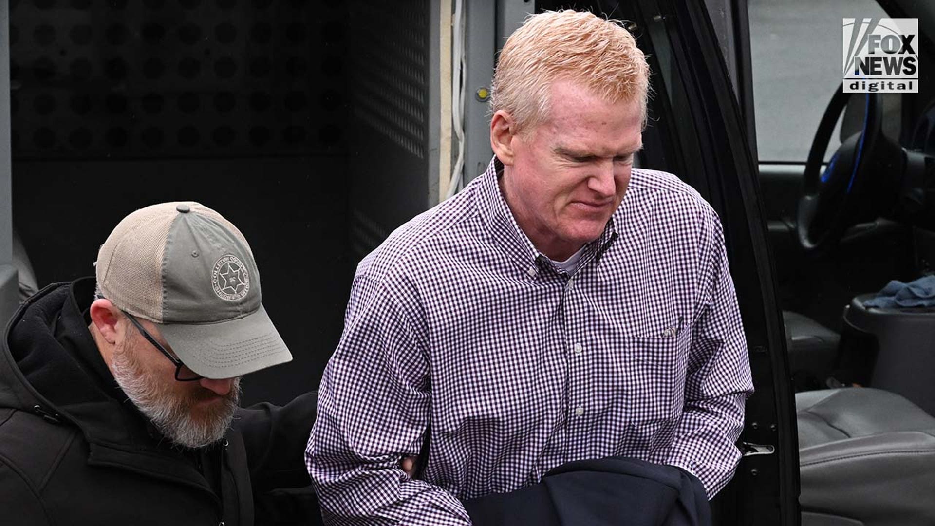 A man wearing a plaid shirt exits a police vehicle surrounded by officers.