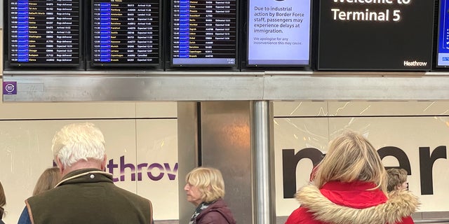 People stand near an arrivals board displaying a message alerting Terminal 5 users to the industrial action by Border Force staff, at Heathrow Airport, near London, on December 23, 2022.  