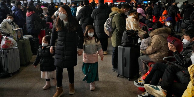 A woman and children wearing face masks walk by masked travelers wait at a departure hall to catch their trains at the West Railway Station in Beijing, Sunday, Jan. 15, 2023.