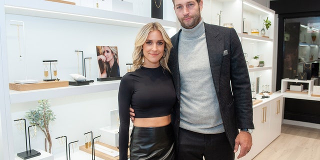 Kristin Cavallari in a long sleeve black crop top shirt and leather bottoms smiles next to Jay Cutler in a black suit jacket and grey turtleneck