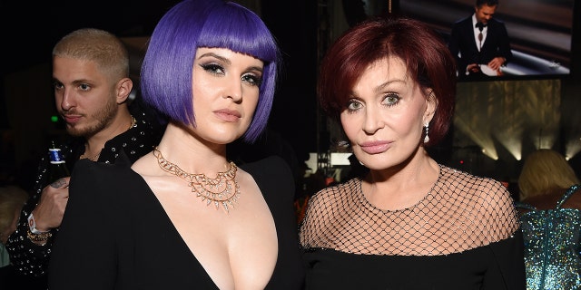 Kelly Osbourne with a short purple bob and deep V-neck black dress smiles for a photo with her mother Sharon in a black gown with a mesh top