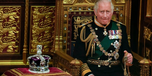 King Charles in green suit adorned with medals and chains sits in a royal chair next to a crown