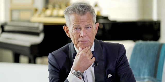 David Foster in a black suit puts his hand on his chin and stares directly at the camera