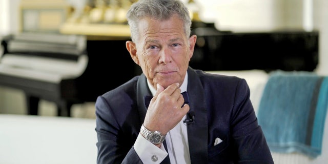 David Foster in a black suit puts his hand on his chin and stares directly at the camera