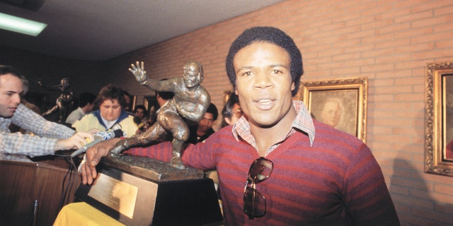 Charles White de la USC, ganador del Trofeo Heisman de 1979, pone su brazo alrededor del mismo trofeo ganado por OJ Simpson en la conferencia de prensa del 12/3.  White, el tercer troyano en ganar el premio, terminó la temporada regular con 1,803 yardas terrestres, la mayor cantidad en la nación.